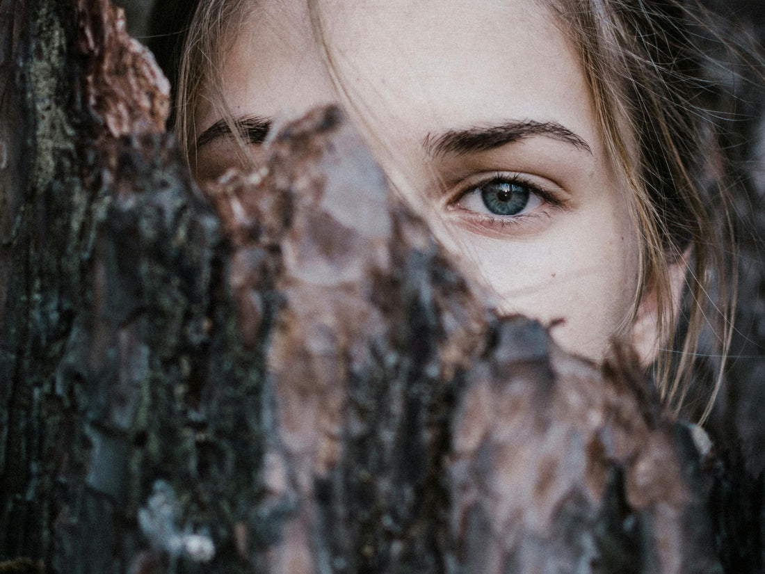 Uma foto de uma mulher por de trás de um tronco quebrado. Apenas é visível uma parte do olhar da mulher
